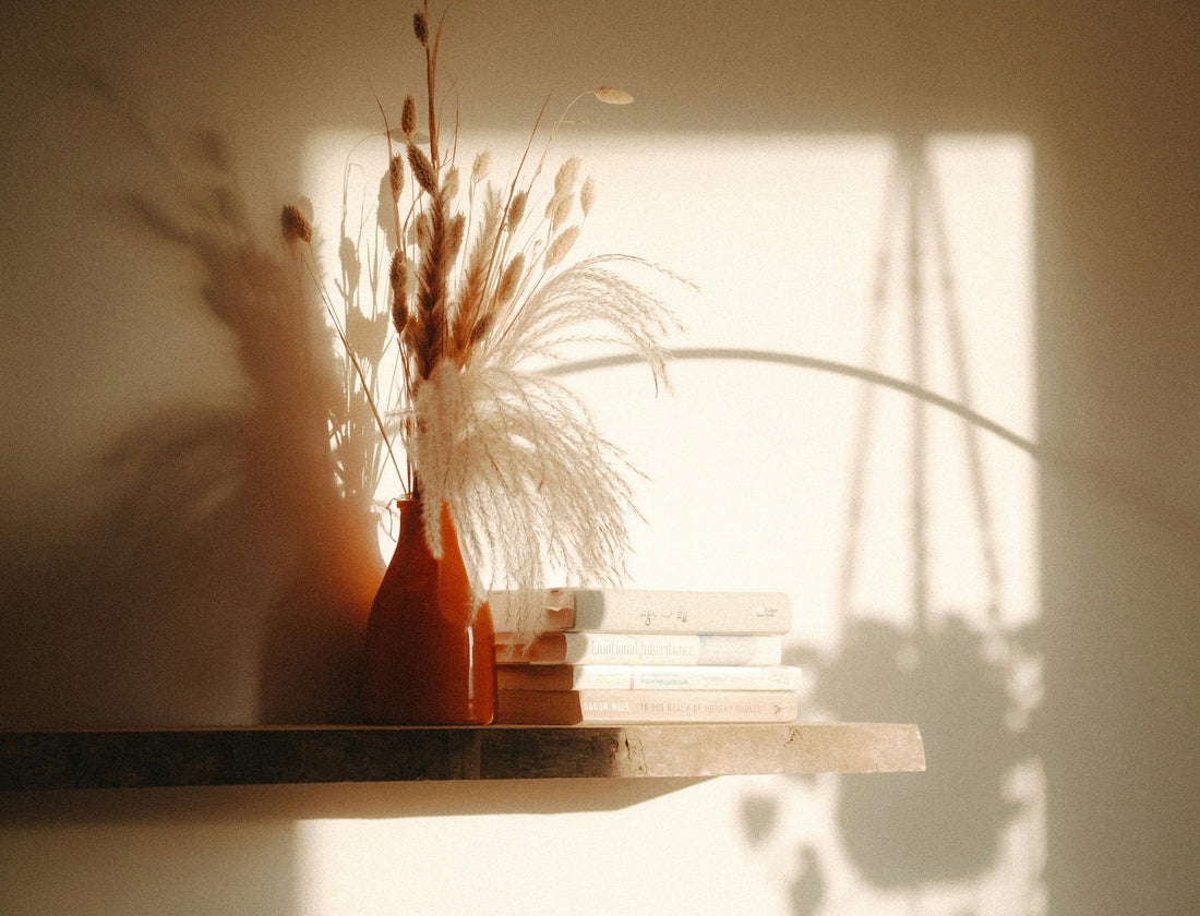 Soft sunlight casting shadows on a shelf with dried flowers in a vase and stacked books