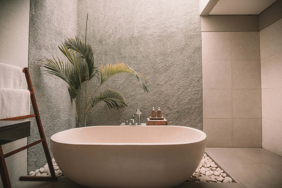 Minimalist spa-like bathroom featuring a freestanding white bathtub surrounded by smooth white stones, a wooden ladder holding white towels, and a potted green plant against a textured gray wall.
