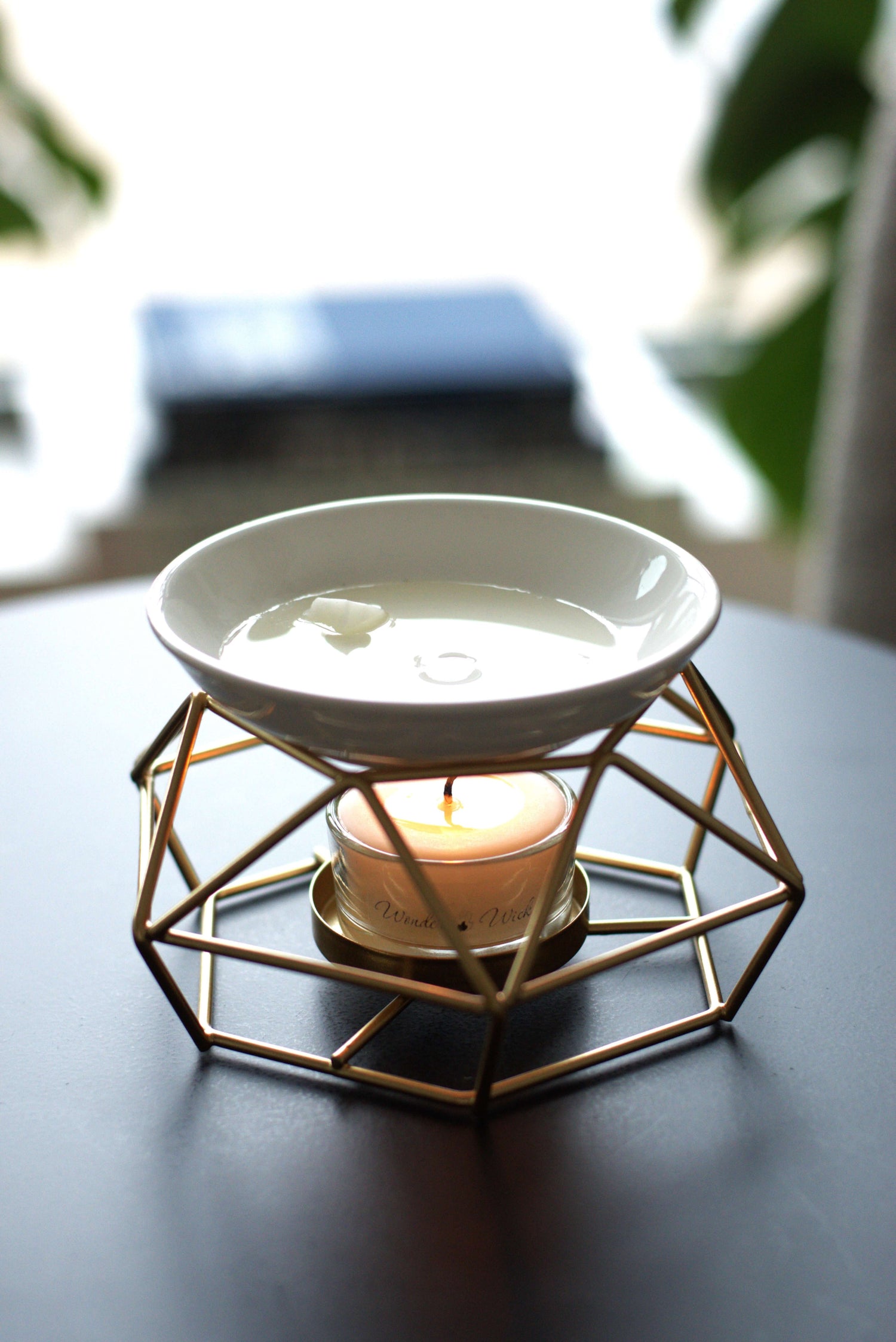 Wonder Wick metal wax melt burner with a Wonder Wick tea light candle, melting an aromatherapy wax melt in the ceramic dish on top. Set on a wooden surface with a cozy, blurred outdoor background.