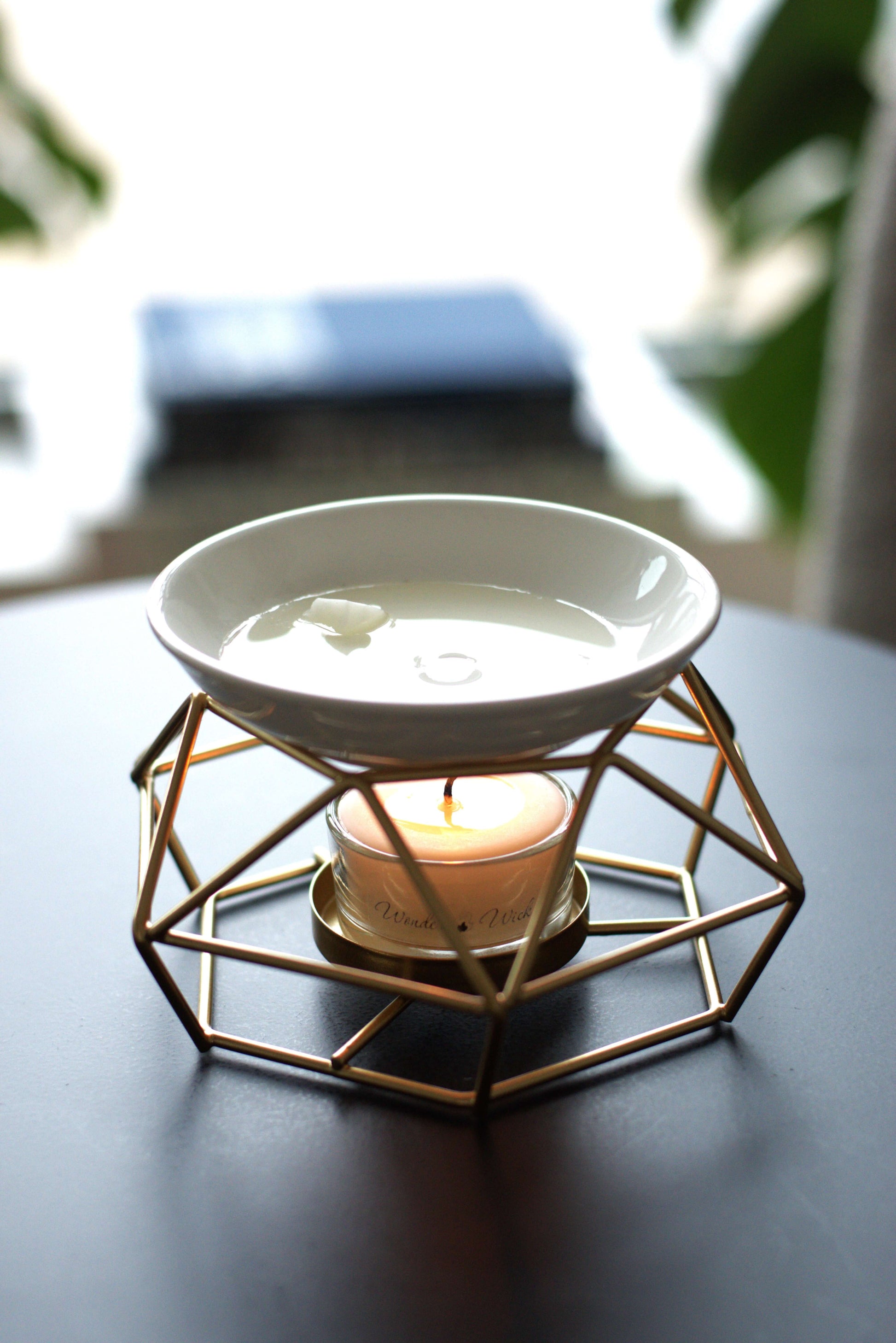 Wonder Wick metal wax melt burner with a Wonder Wick tea light candle, melting an aromatherapy wax melt in the ceramic dish on top. Set on a wooden surface with a cozy, blurred outdoor background.
