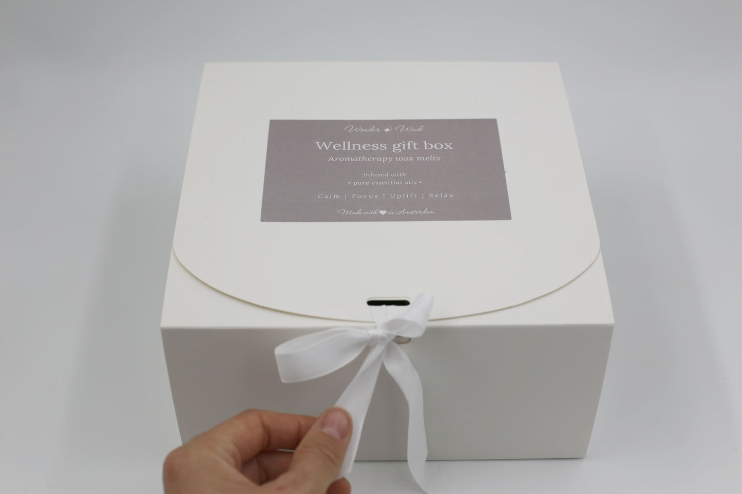 Hand unties a white ribbon on Wonder Wick’s Wellness Gift Box for essential oil wax melts. The gray label reads 'Wellness gift box - Aromatherapy wax melts' with calming benefits 'Calm, Focus, Uplift, Relax,' ideal for gifting.
