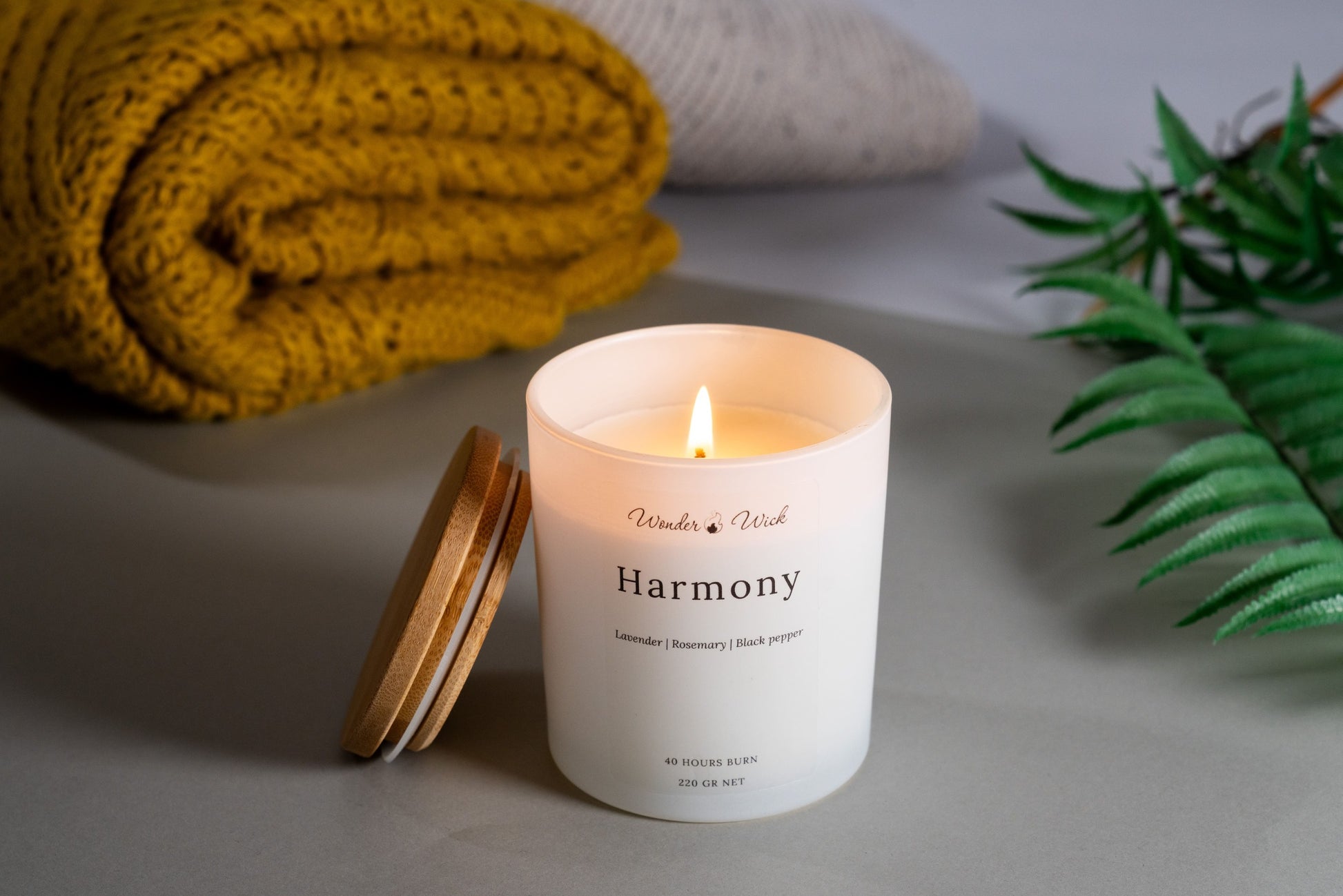 A lit 'Harmony' aromatherapy candle, crafted from coconut and rapeseed wax mixed with pure essential oils of lavender, rosemary, and black pepper. The relaxing candle, emitting a gentle glow, is placed next to a yellow knitted throw and lush green plant leaves, enhancing a serene and balanced environment.