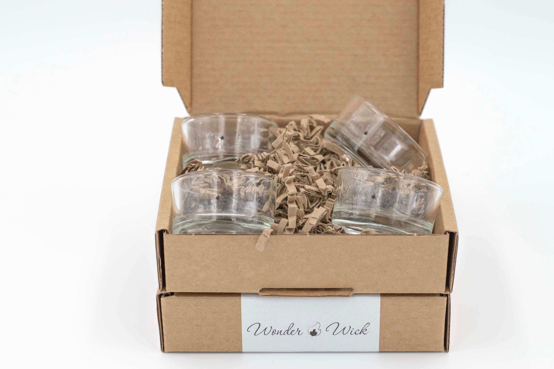 Four glass tea light holders packaged in a cardboard box with shredded paper for protection. The box displays the Wonder Wick logo.