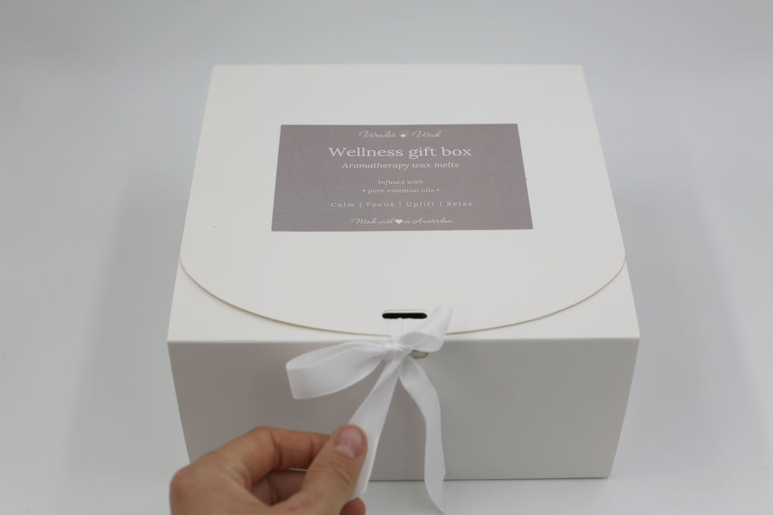 White wellness gift box tied with a white ribbon, labeled 'Wellness Gift Box' containing aromatherapy wax melts, tealight candles, and other wellness items, with a hand reaching to untie the ribbon.