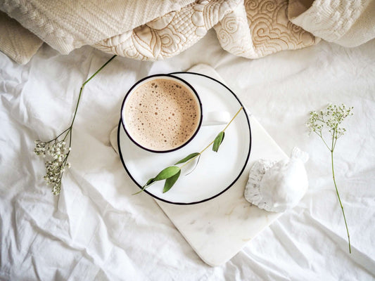 Flat lay of cosy winter essentials featuring a warm cup of frothy coffee, soft blankets, and delicate greenery, creating a relaxing and calming atmosphere.