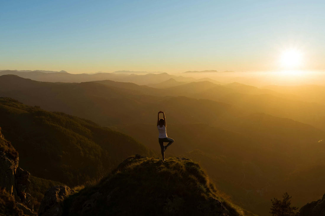 Woman practicing yoga on a mountain peak at sunrise, symbolizing the calm and rejuvenating benefits of yoga in nature.