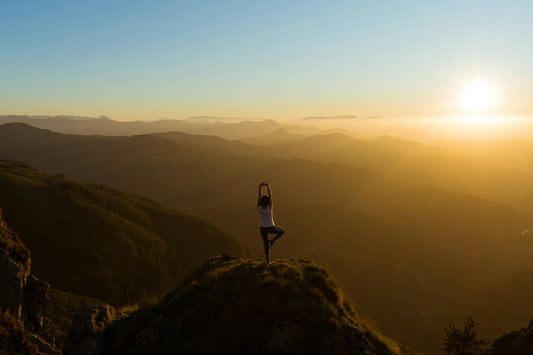 Woman practicing yoga on a mountain peak at sunrise, symbolizing the calm and rejuvenating benefits of yoga in nature.