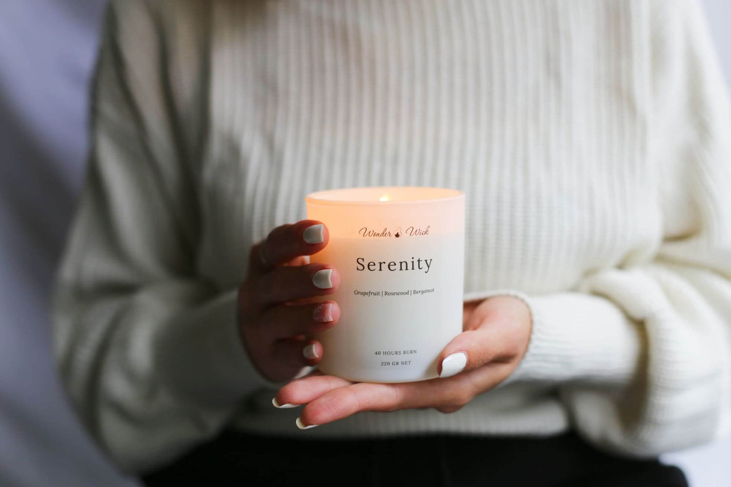 Person holding a lit Serenity aromatherapy candle by Wonder Wick, featuring a white container labeled 'Serenity' with grapefruit, rosewood, and bergamot essential oils, designed for relaxation and mindfulness.