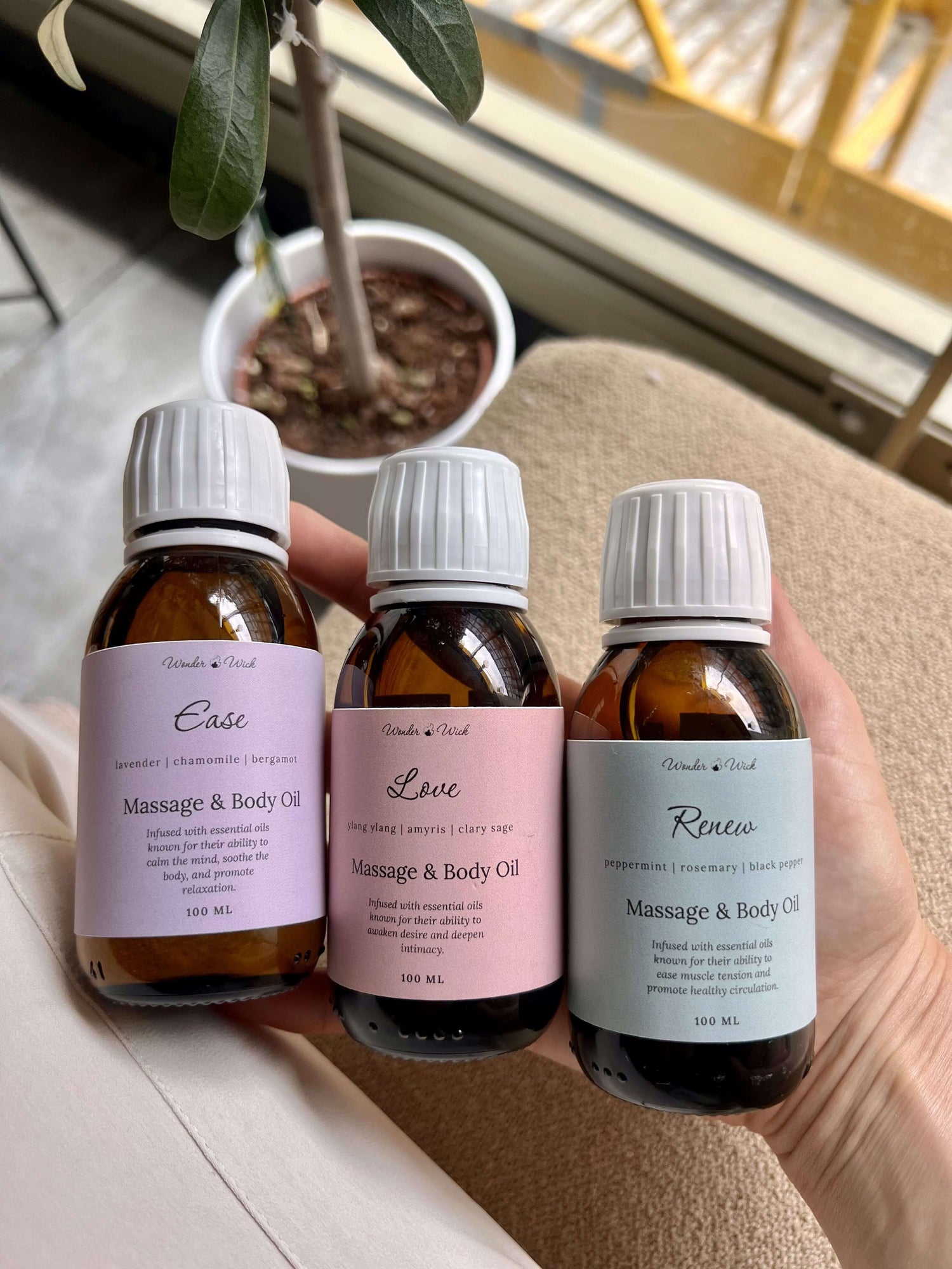 Three bottles of aromatherapy massage oils held in hand. Each bottle is labeled: Ease (lavender, chamomile, bergamot), Love (ylang ylang, amyris, clary sage), and Renew (peppermint, rosemary, black pepper).
