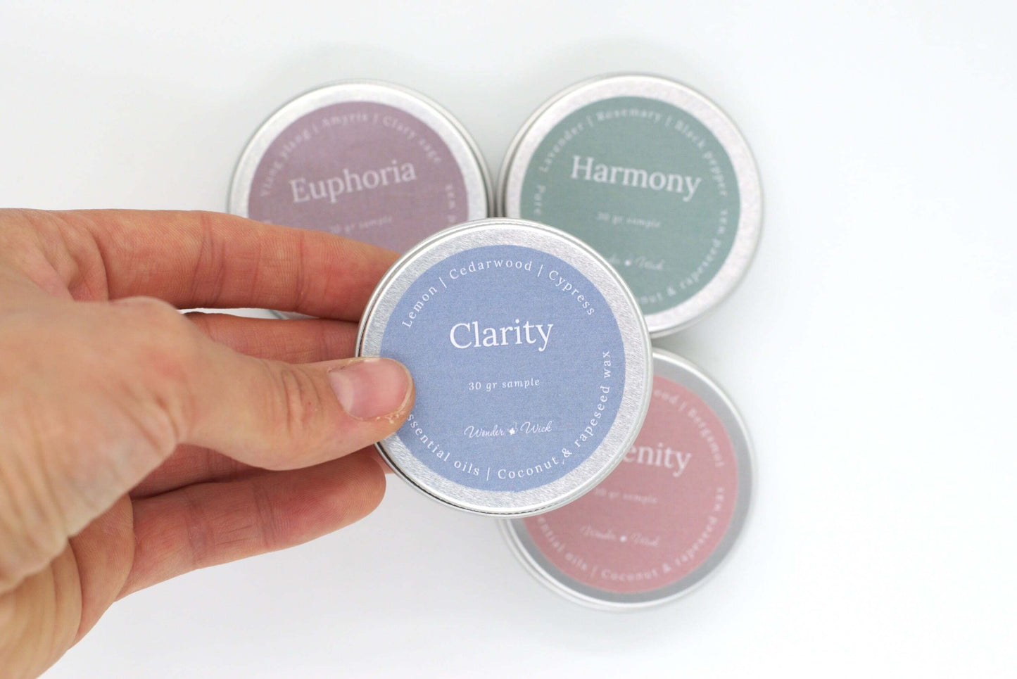 Close-up of a hand holding a tin sample of Clarity aromatherapy candle, featuring essential oils of lemon, cedarwood, and cypress. The blue label displays 30g sample size and notes of pure essential oils in coconut and rapeseed wax