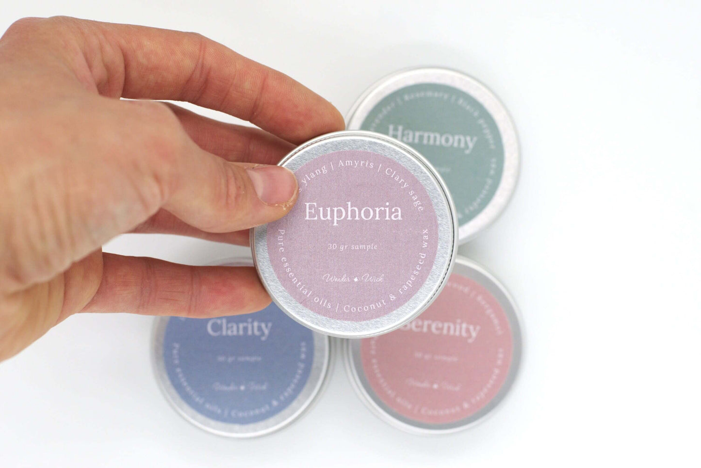 Close-up of a hand holding a tin sample of Euphoria aromatherapy candle, labeled with the scent of ylang ylang, amyris, and clary sage. The pink-purple label highlights the 30g sample size and the use of pure essential oils blended with coconut and rapese