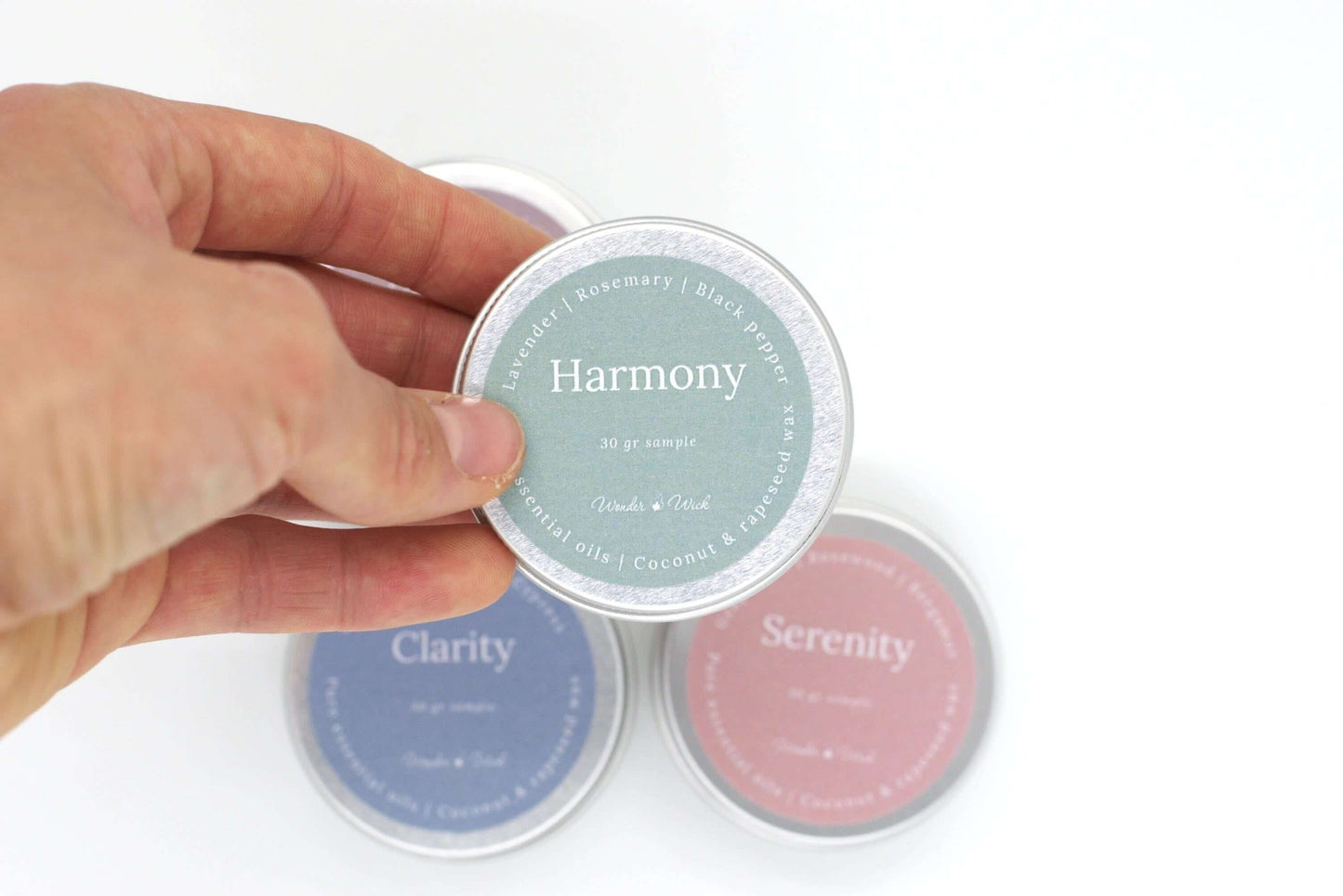 Close-up of a hand holding a tin sample of Harmony aromatherapy candle, scented with lavender, rosemary, and black pepper. The green label indicates a 30g sample size with a blend of pure essential oils and coconut and rapeseed wax.