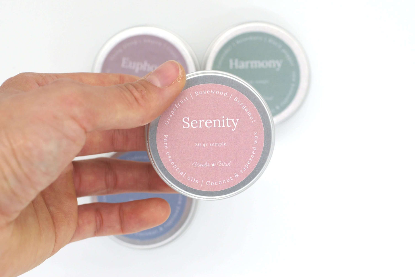 Close-up of a hand holding a tin sample of Serenity aromatherapy candle, scented with grapefruit, rosewood, and bergamot. The pink label shows 30g sample size, highlighting natural ingredients with pure essential oils in coconut and rapeseed wax.