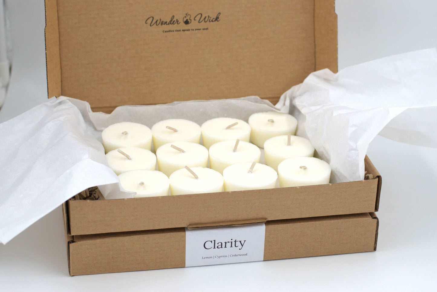 Open cardboard box containing twelve Clarity essential oil tealight refills by Wonder Wick, made with lemon, cypress and cedarwood and neatly arranged and wrapped in eco-friendly tissue paper. 