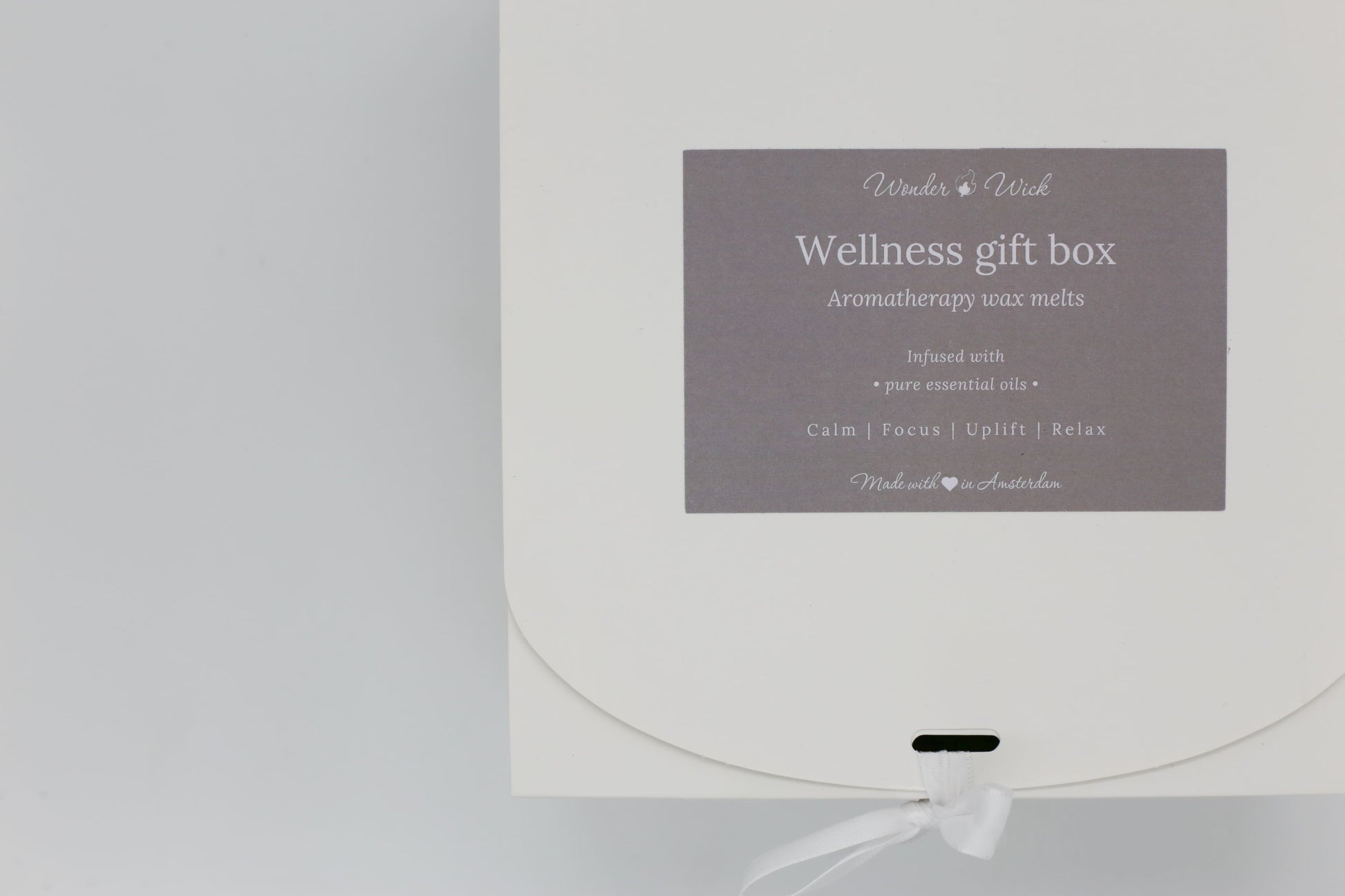 Close-up of Wonder Wick Wellness Gift Box for aromatherapy wax melts, featuring a gray label with 'Wellness gift box - Aromatherapy wax melts,' benefits like 'Calm, Focus, Uplift, Relax,' and 'Made with love in Amsterdam,' tied with a white ribbon.