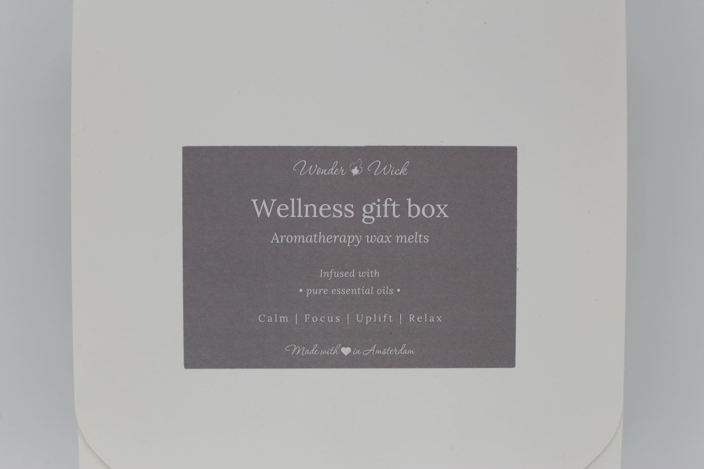 Detailed view of Wonder Wick Wellness Gift Box label for aromatherapy wax melts. Label reads 'Wellness gift box - Aromatherapy wax melts,' with benefits 'Calm, Focus, Uplift, Relax,' and 'Made with love in Amsterdam' in soft gray.