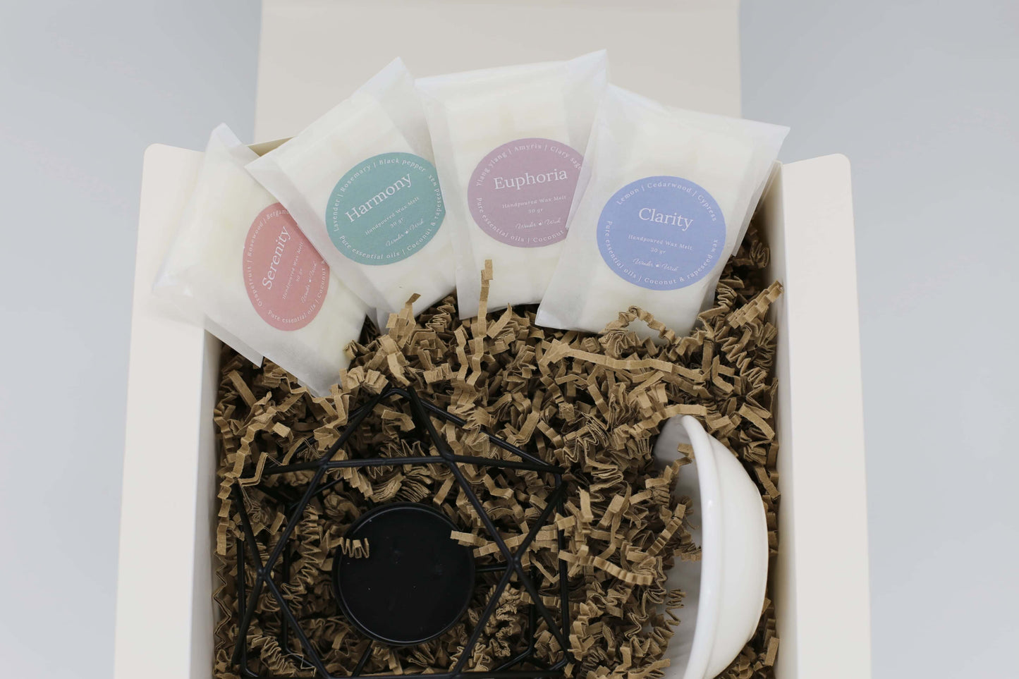 Wonder Wick Wellness Gift Box opened to show four wrapped wax melts: Serenity, Harmony, Euphoria, and Clarity. Each wax melt is labeled with ingredients, nestled in eco-friendly packing paper, with a oil burner with ceramic dish.