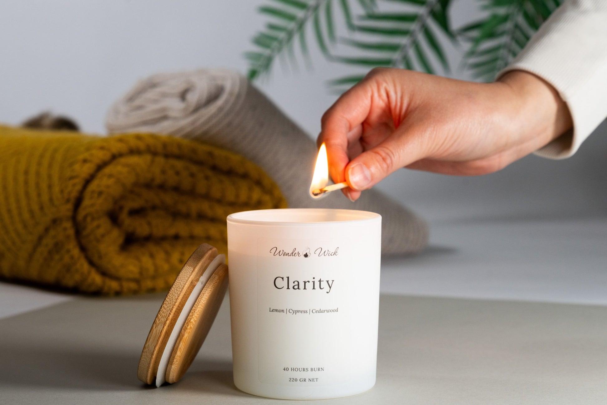 A hand lighting a 'Clarity' aromatherapy candle with a match. This white candle, made from a coconut and rapeseed wax base and infused with lemon, cypress, and cedarwood essential oils, is positioned near a vibrant yellow knitted throw and greenery, promoting a clear and refreshed ambiance.