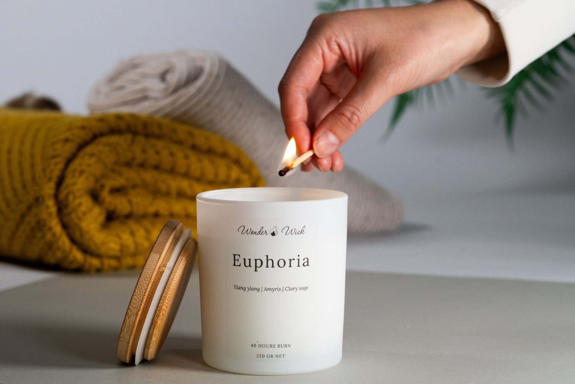 A hand lighting a white 'Euphoria' aromatherapy candle made from a blend of coconut and rapeseed wax, enriched with essential oils of ylang ylang, amaryllis, and clary sage. The candle is set against a backdrop of a cozy yellow knitted throw and a grey sock, suggesting a warm, inviting atmosphere.