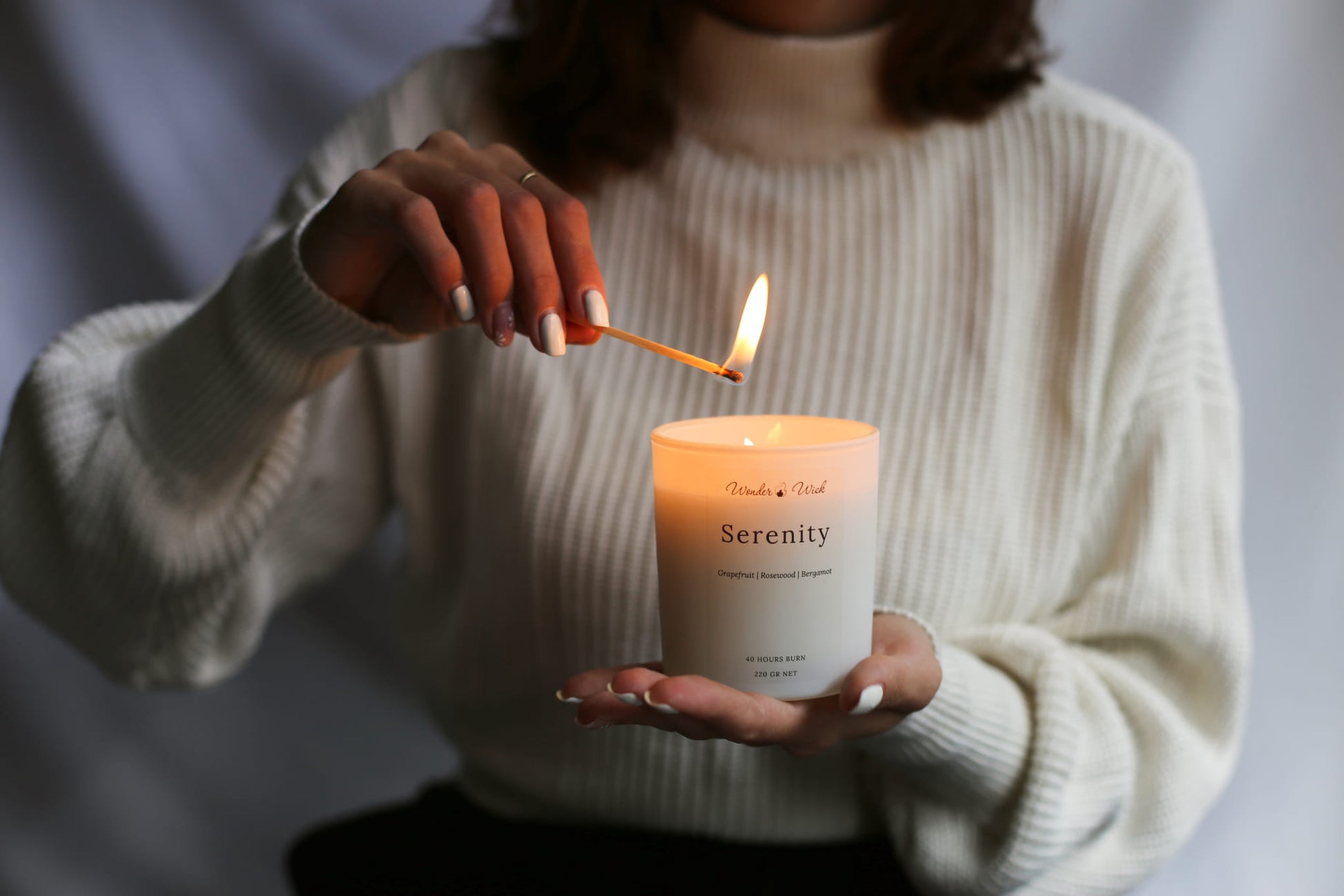 A person in a white sweater lights the Wonder Wick "Serenity" candle, featuring a soothing blend of grapefruit, rosewood, and bergamot essential oils. The candle offers a 40-hour burn time and is designed to promote relaxation and mood enhancement through aromatherapy.