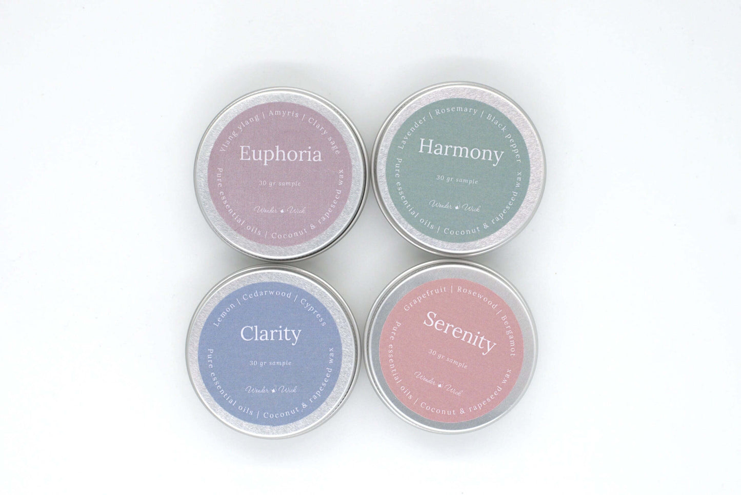 Overhead view of four aromatherapy candle samples in tin containers, featuring Clarity, Euphoria, Harmony, and Serenity scents. Labels on each tin display respective essential oil blends: lemon, cedarwood, and cypress for Clarity; ylang ylang, amyris, and