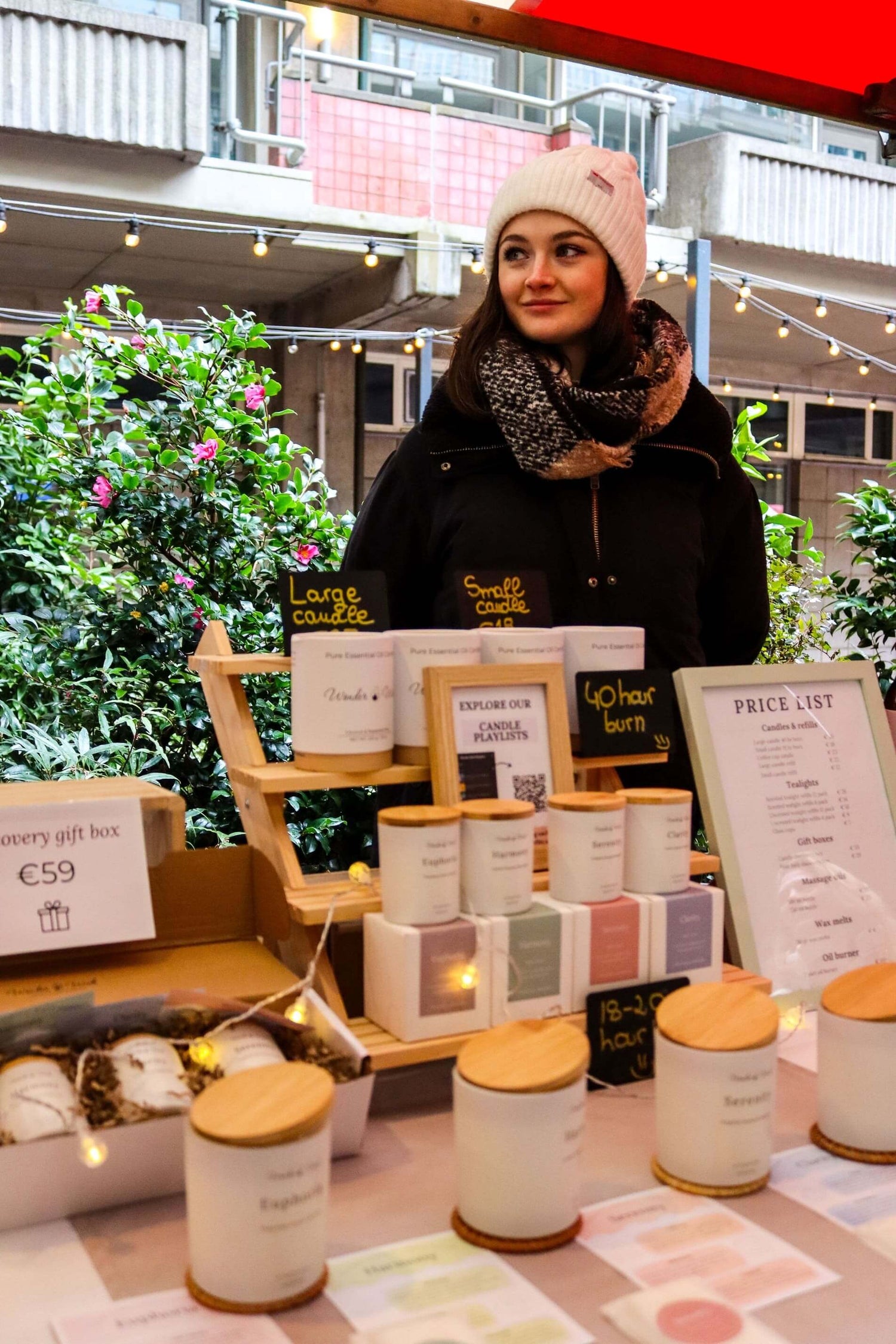 A woman in a white beanie and black coat stands behind a market stall displaying Wonder Wick essential oil candles in glass jars, candle gift boxes, and a price list. The stand is decorated with string lights, and chalkboard signs label the candles as “large” and “small.” A green plant and urban buildings are visible in the background.